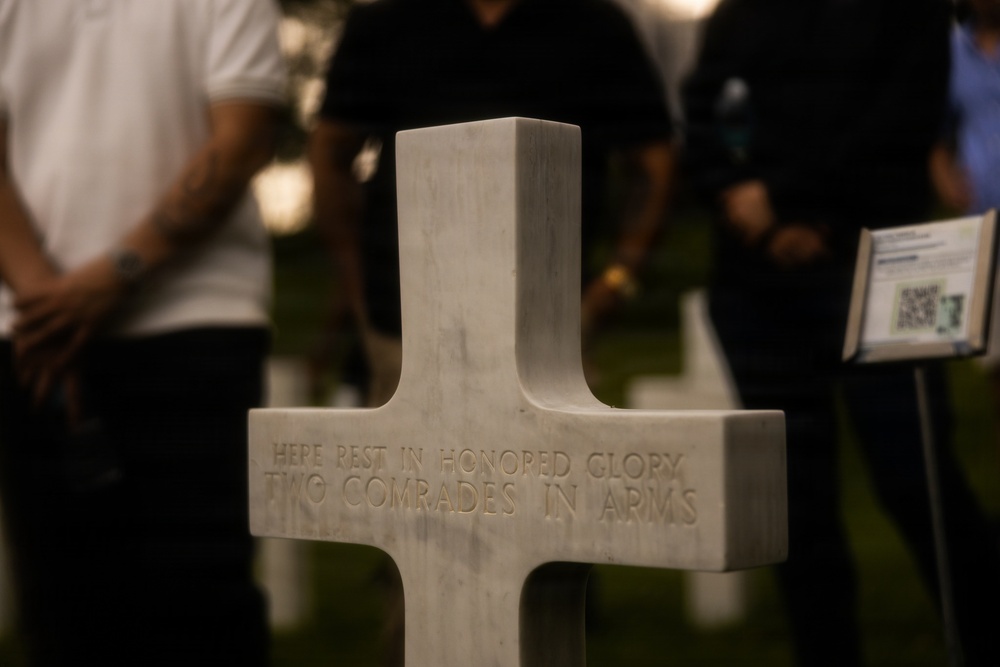 A white marble military cross gravestone is in sharp focus in the foreground, inscribed with 'HERE REST IN HONORED GLORY TWO COMRADES IN ARMS.' The setting appears to be a military cemetery, likely an American war cemetery, during an evening or nighttime commemorative event. Several people are visible in the blurred background, along with what appears to be an informational display stand with a QR code.
