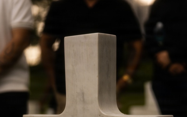 A white marble military cross gravestone is in sharp focus in the foreground, inscribed with 'HERE REST IN HONORED GLORY TWO COMRADES IN ARMS.' The setting appears to be a military cemetery, likely an American war cemetery, during an evening or nighttime commemorative event. Several people are visible in the blurred background, along with what appears to be an informational display stand with a QR code.