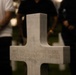 A white marble military cross gravestone is in sharp focus in the foreground, inscribed with 'HERE REST IN HONORED GLORY TWO COMRADES IN ARMS.' The setting appears to be a military cemetery, likely an American war cemetery, during an evening or nighttime commemorative event. Several people are visible in the blurred background, along with what appears to be an informational display stand with a QR code.