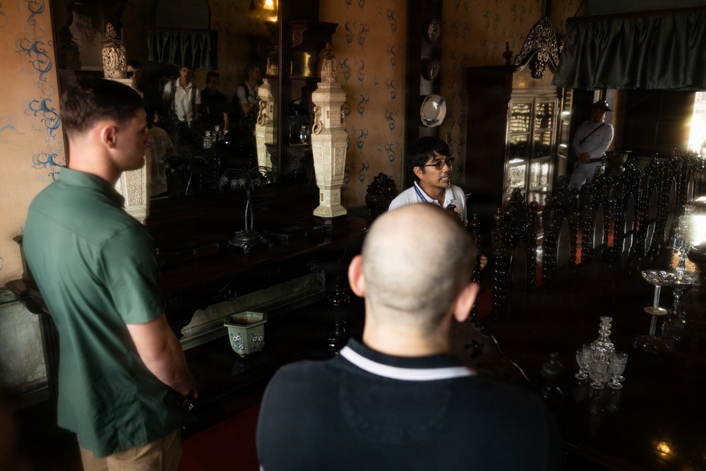 A group of people appear to be on a guided tour inside an ornate, historic interior space filled with antique furniture and decorative objects. A guide wearing glasses and a white shirt appears to be speaking to visitors, including a man in a green shirt and a bald man in a dark polo. The room features large mirrors, dark wooden furniture, crystal glassware, and elaborate wall decorations.