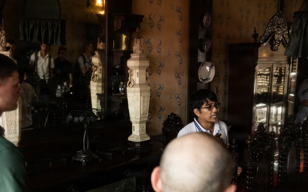 A group of people appear to be on a guided tour inside an ornate, historic interior space filled with antique furniture and decorative objects. A guide wearing glasses and a white shirt appears to be speaking to visitors, including a man in a green shirt and a bald man in a dark polo. The room features large mirrors, dark wooden furniture, crystal glassware, and elaborate wall decorations.