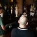 A group of people appear to be on a guided tour inside an ornate, historic interior space filled with antique furniture and decorative objects. A guide wearing glasses and a white shirt appears to be speaking to visitors, including a man in a green shirt and a bald man in a dark polo. The room features large mirrors, dark wooden furniture, crystal glassware, and elaborate wall decorations.
