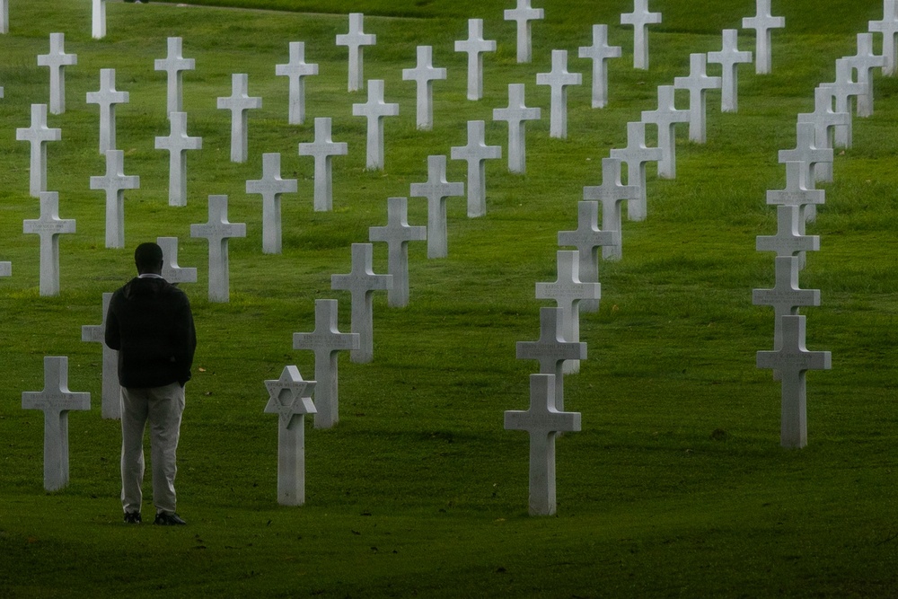 A solitary figure stands among rows of white marble grave markers at what appears to be an American military cemetery, likely in Europe. The cemetery features predominantly Latin crosses with at least one Star of David marker visible, consistent with U.S. military burial grounds such as the Normandy American Cemetery. The scene conveys quiet reflection and remembrance amid a vast, well-maintained green landscape.
