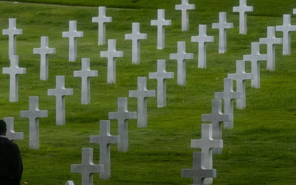 A solitary figure stands among rows of white marble grave markers at what appears to be an American military cemetery, likely in Europe. The cemetery features predominantly Latin crosses with at least one Star of David marker visible, consistent with U.S. military burial grounds such as the Normandy American Cemetery. The scene conveys quiet reflection and remembrance amid a vast, well-maintained green landscape.