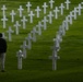 A solitary figure stands among rows of white marble grave markers at what appears to be an American military cemetery, likely in Europe. The cemetery features predominantly Latin crosses with at least one Star of David marker visible, consistent with U.S. military burial grounds such as the Normandy American Cemetery. The scene conveys quiet reflection and remembrance amid a vast, well-maintained green landscape.