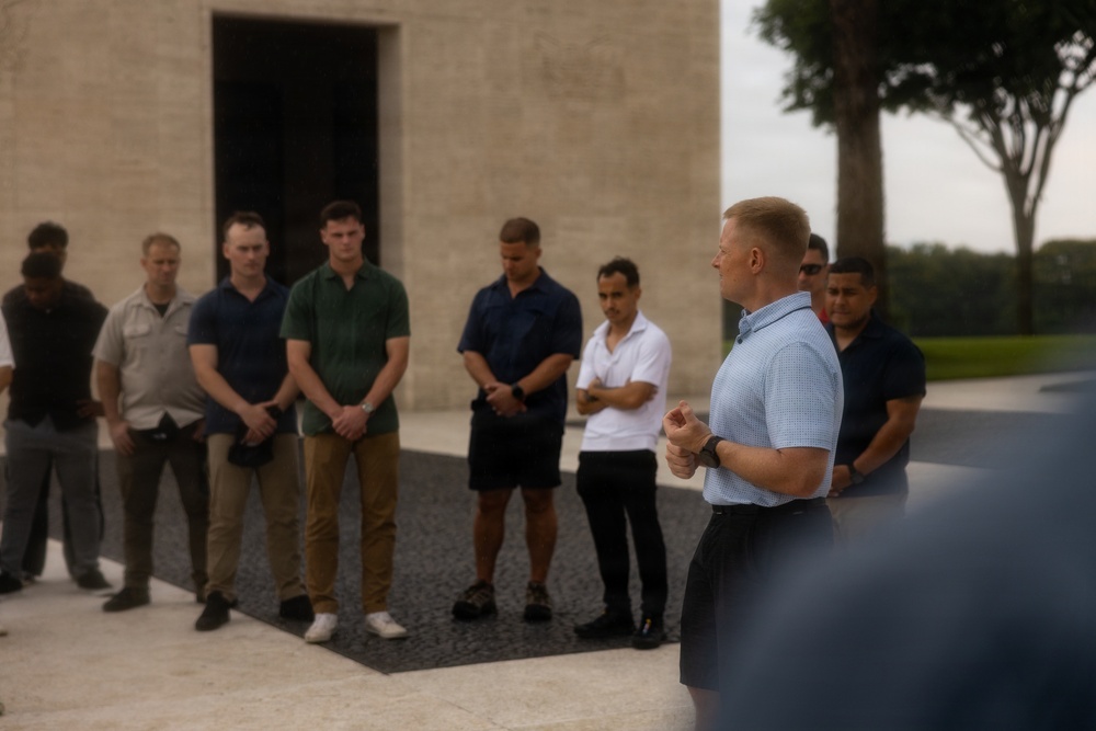 A man in a light blue polo shirt appears to be addressing a group of approximately 8-10 individuals standing in a line outdoors. The setting appears to be a formal or institutional outdoor space, with a large stone or concrete building visible in the background. The group is listening attentively, with several individuals looking downward in a respectful or contemplative posture.