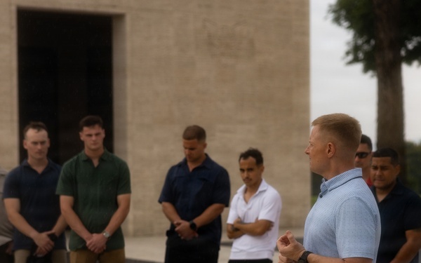 A man in a light blue polo shirt appears to be addressing a group of approximately 8-10 individuals standing in a line outdoors. The setting appears to be a formal or institutional outdoor space, with a large stone or concrete building visible in the background. The group is listening attentively, with several individuals looking downward in a respectful or contemplative posture.