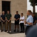 A man in a light blue polo shirt appears to be addressing a group of approximately 8-10 individuals standing in a line outdoors. The setting appears to be a formal or institutional outdoor space, with a large stone or concrete building visible in the background. The group is listening attentively, with several individuals looking downward in a respectful or contemplative posture.