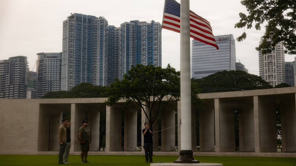 A person is raising or lowering an American flag on a tall flagpole at what appears to be a military memorial or cemetery, identifiable by the colonnaded stone wall structure in the background. Several individuals stand nearby observing the ceremony. The scene is set against a backdrop of modern high-rise buildings, suggesting an urban Asian city location.