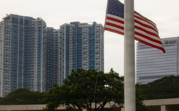 A person is raising or lowering an American flag on a tall flagpole at what appears to be a military memorial or cemetery, identifiable by the colonnaded stone wall structure in the background. Several individuals stand nearby observing the ceremony. The scene is set against a backdrop of modern high-rise buildings, suggesting an urban Asian city location.