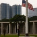 A person is raising or lowering an American flag on a tall flagpole at what appears to be a military memorial or cemetery, identifiable by the colonnaded stone wall structure in the background. Several individuals stand nearby observing the ceremony. The scene is set against a backdrop of modern high-rise buildings, suggesting an urban Asian city location.
