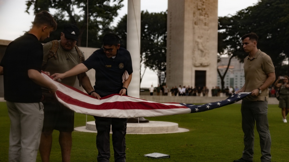 Four individuals are carefully folding an American flag on a grass lawn in front of a large monument or memorial structure. The scene appears to be a formal flag retirement or lowering ceremony, likely at a military or national memorial site. A crowd of observers is visible in the background near the monument.