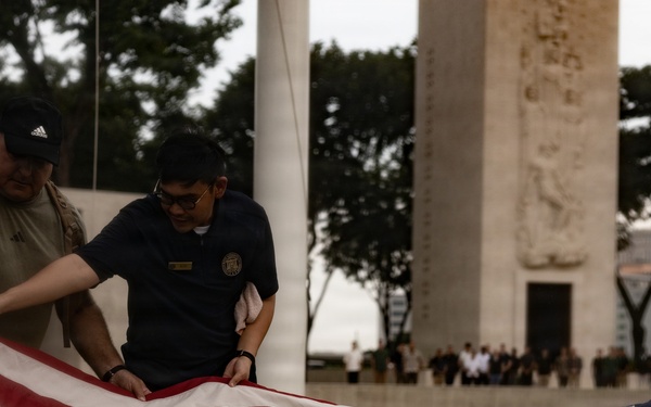 Four individuals are carefully folding an American flag on a grass lawn in front of a large monument or memorial structure. The scene appears to be a formal flag retirement or lowering ceremony, likely at a military or national memorial site. A crowd of observers is visible in the background near the monument.