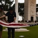 Four individuals are carefully folding an American flag on a grass lawn in front of a large monument or memorial structure. The scene appears to be a formal flag retirement or lowering ceremony, likely at a military or national memorial site. A crowd of observers is visible in the background near the monument.