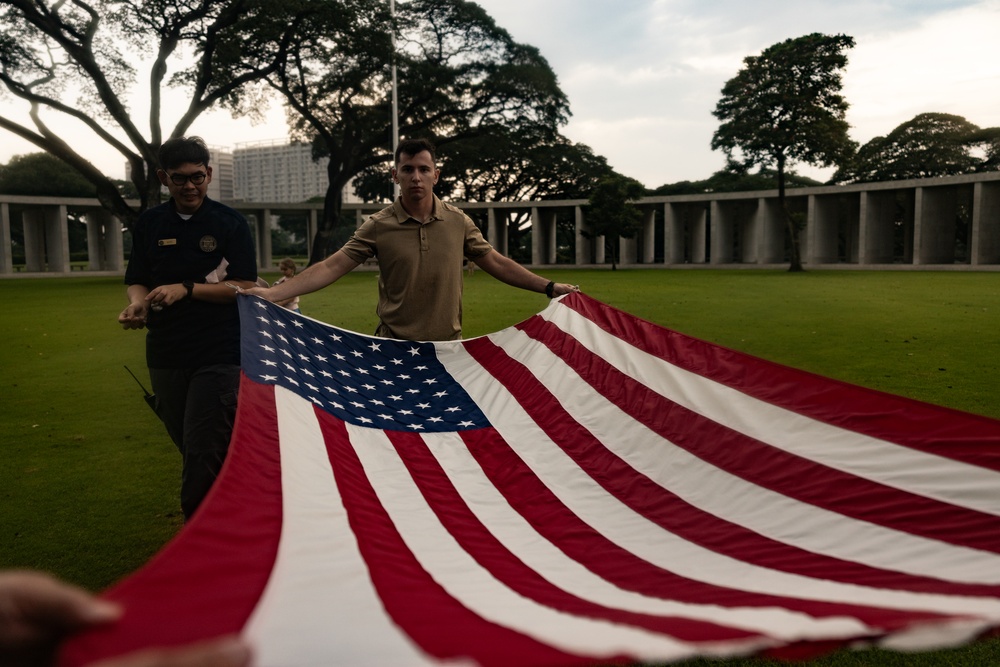 Two individuals are folding or unfolding a large American flag on a well-maintained green lawn. The setting appears to be a formal memorial or ceremonial grounds, identifiable by the colonnaded structure and large trees in the background. The scene suggests a flag ceremony, possibly at a military cemetery or memorial site.