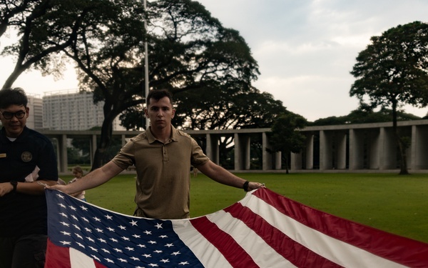 Two individuals are folding or unfolding a large American flag on a well-maintained green lawn. The setting appears to be a formal memorial or ceremonial grounds, identifiable by the colonnaded structure and large trees in the background. The scene suggests a flag ceremony, possibly at a military cemetery or memorial site.
