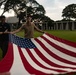 Two individuals are folding or unfolding a large American flag on a well-maintained green lawn. The setting appears to be a formal memorial or ceremonial grounds, identifiable by the colonnaded structure and large trees in the background. The scene suggests a flag ceremony, possibly at a military cemetery or memorial site.