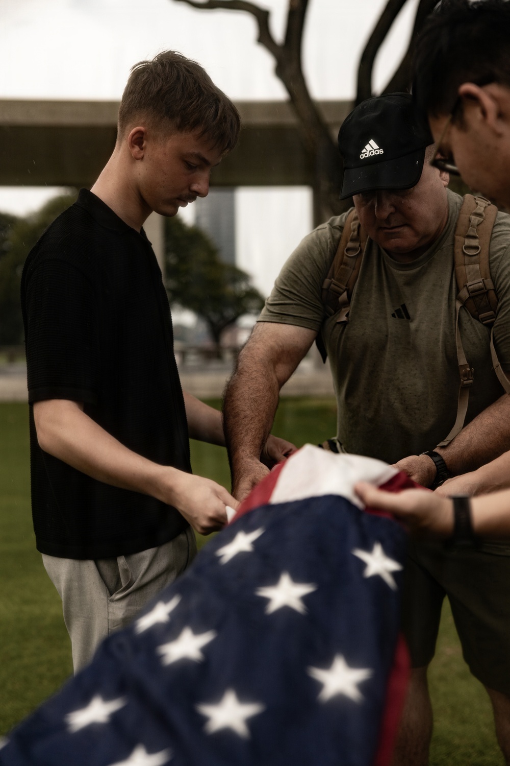 Three individuals are gathered outdoors carefully folding or handling an American flag. The group includes a young man in a black shirt, an older man wearing an Adidas cap and olive t-shirt with a tactical backpack, and a third person partially visible on the right. The setting appears to be a grassy outdoor area with trees and urban structures in the background.