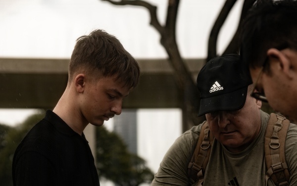 Three individuals are gathered outdoors carefully folding or handling an American flag. The group includes a young man in a black shirt, an older man wearing an Adidas cap and olive t-shirt with a tactical backpack, and a third person partially visible on the right. The setting appears to be a grassy outdoor area with trees and urban structures in the background.