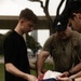 Three individuals are gathered outdoors carefully folding or handling an American flag. The group includes a young man in a black shirt, an older man wearing an Adidas cap and olive t-shirt with a tactical backpack, and a third person partially visible on the right. The setting appears to be a grassy outdoor area with trees and urban structures in the background.