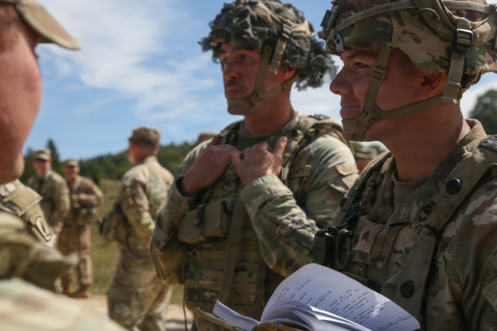 A group of military personnel in U.S. Army camouflage (OCP/Multicam) uniforms and combat helmets are engaged in what appears to be a briefing or training exercise outdoors. One soldier in the foreground is holding an open notebook with handwritten notes visible. The setting appears to be a field training environment with trees and open terrain in the background.