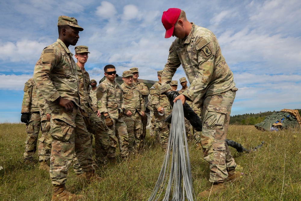 A group of U.S. Army soldiers in OCP (Operational Camouflage Pattern) uniforms are gathered in an open field during what appears to be a training exercise. One soldier wearing a distinctive red baseball cap is demonstrating the handling or inspection of parachute suspension lines or similar cordage to the assembled group. Military equipment, including what appears to be a parachute or tent structure, is visible in the background.