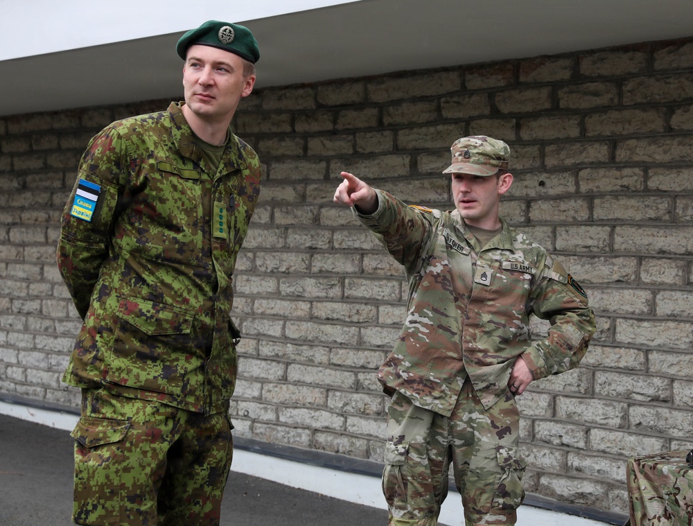 Two military personnel are engaged in conversation outside a stone/brick building. The soldier on the left wears an Estonian military uniform with a green beret and an Estonian flag patch along with a 'Слава Україні' (Glory to Ukraine) patch. The soldier on the right wears a U.S. Army MultiCam uniform and is pointing in a direction while speaking.