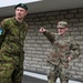 Two military personnel are engaged in conversation outside a stone/brick building. The soldier on the left wears an Estonian military uniform with a green beret and an Estonian flag patch along with a 'Слава Україні' (Glory to Ukraine) patch. The soldier on the right wears a U.S. Army MultiCam uniform and is pointing in a direction while speaking.