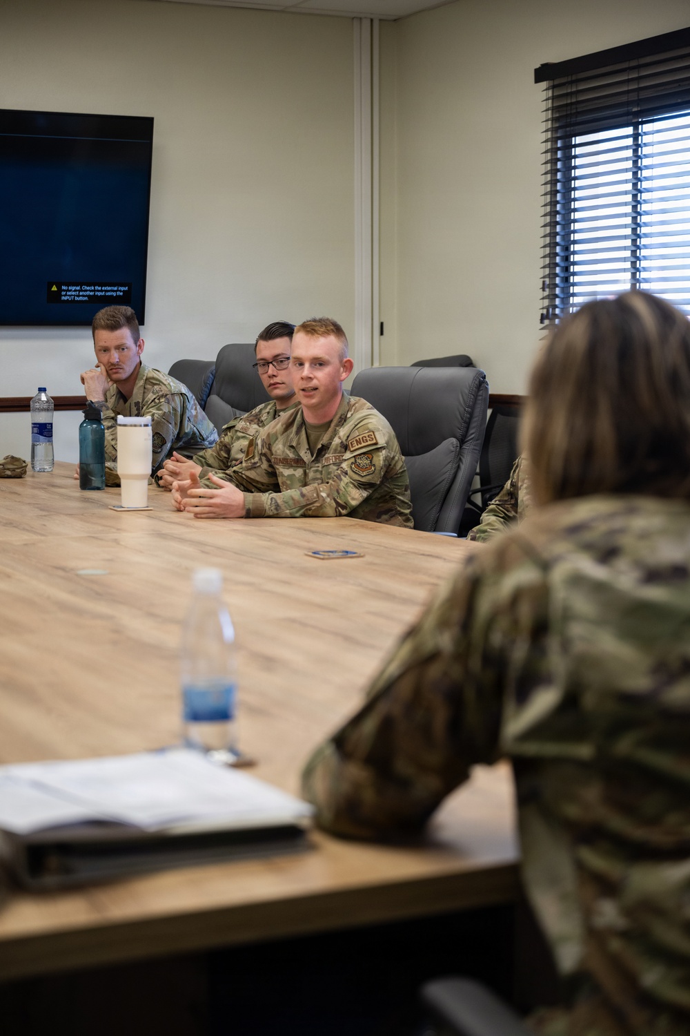 A group of military personnel in OCP (Operational Camouflage Pattern) uniforms are seated around a conference table in what appears to be a briefing or meeting room. One airman wearing an 'ENGS' (Engineers) patch appears to be speaking or presenting to others at the table. A wall-mounted display screen in the background shows a 'No signal' message.