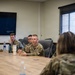 A group of military personnel in OCP (Operational Camouflage Pattern) uniforms are seated around a conference table in what appears to be a briefing or meeting room. One airman wearing an 'ENGS' (Engineers) patch appears to be speaking or presenting to others at the table. A wall-mounted display screen in the background shows a 'No signal' message.