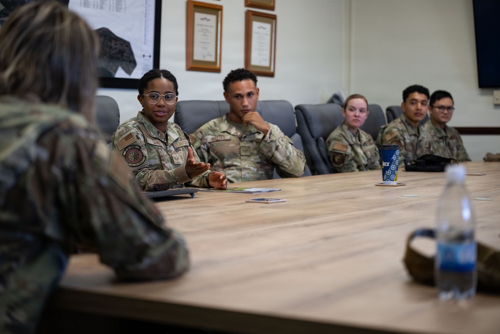 A group of military personnel in OCP (Operational Camouflage Pattern) uniforms are seated around a conference table in what appears to be a briefing or meeting room. A female service member wearing glasses is actively speaking and gesturing with her hand, appearing to lead or contribute to the discussion. The setting includes framed documents and maps on the wall, suggesting an official military workspace.