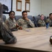 A group of military personnel in OCP (Operational Camouflage Pattern) uniforms are seated around a conference table in what appears to be a briefing or meeting room. A female service member wearing glasses is actively speaking and gesturing with her hand, appearing to lead or contribute to the discussion. The setting includes framed documents and maps on the wall, suggesting an official military workspace.