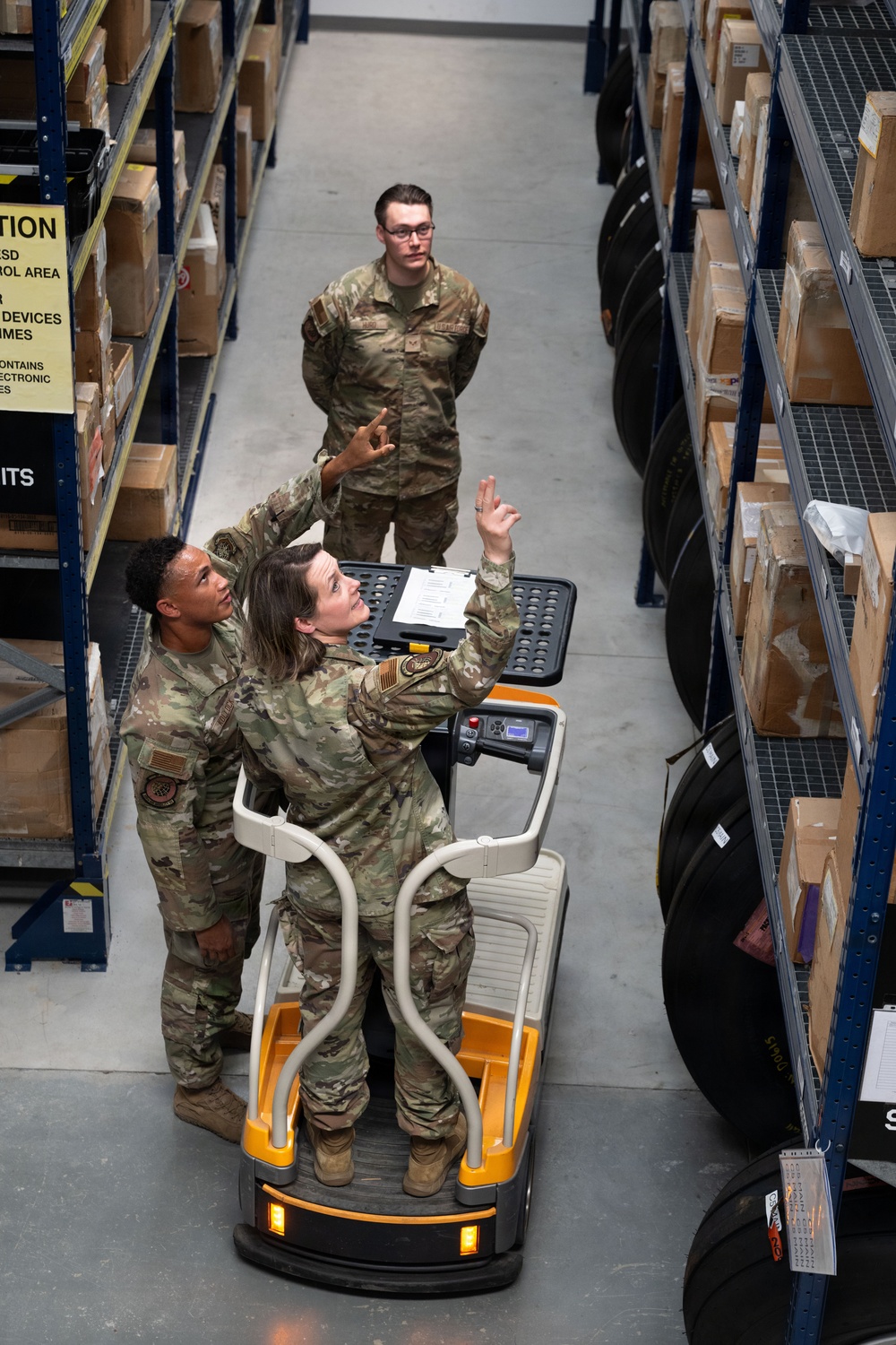 Three military personnel in OCP (Operational Camouflage Pattern) uniforms are working in a warehouse or supply storage facility. A female service member operating a motorized warehouse vehicle is pointing upward toward shelving while holding a clipboard, apparently directing or instructing two other personnel. The setting appears to be a military logistics or supply depot with tall metal shelving units stocked with boxed inventory.