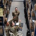 Three military personnel in OCP (Operational Camouflage Pattern) uniforms are working in a warehouse or supply storage facility. A female service member operating a motorized warehouse vehicle is pointing upward toward shelving while holding a clipboard, apparently directing or instructing two other personnel. The setting appears to be a military logistics or supply depot with tall metal shelving units stocked with boxed inventory.