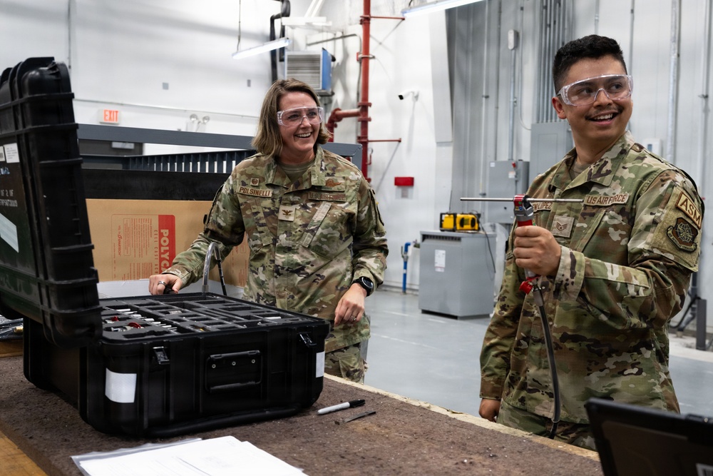 Two U.S. Air Force personnel in OCP (Operational Camouflage Pattern) uniforms are working in what appears to be a maintenance or technical workshop facility. Both individuals are wearing safety goggles and appear to be in good spirits, smiling while handling equipment. The person on the left, identified by nametape as 'POLSINELLI,' is opening a hard-shell equipment case, while the person on the right holds a tool.