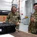 Two U.S. Air Force personnel in OCP (Operational Camouflage Pattern) uniforms are working in what appears to be a maintenance or technical workshop facility. Both individuals are wearing safety goggles and appear to be in good spirits, smiling while handling equipment. The person on the left, identified by nametape as 'POLSINELLI,' is opening a hard-shell equipment case, while the person on the right holds a tool.