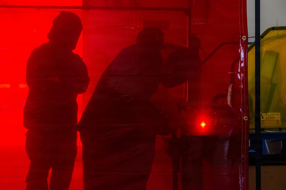 The image shows silhouettes of multiple people visible through a translucent red barrier or curtain, creating a dramatic high-contrast visual effect. A bright red point light source is visible near the center-lower area of the frame. To the right, a yellow barrier or sign with partial text reading 'HOT' is visible beyond the red curtain's edge.