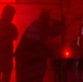 The image shows silhouettes of multiple people visible through a translucent red barrier or curtain, creating a dramatic high-contrast visual effect. A bright red point light source is visible near the center-lower area of the frame. To the right, a yellow barrier or sign with partial text reading 'HOT' is visible beyond the red curtain's edge.