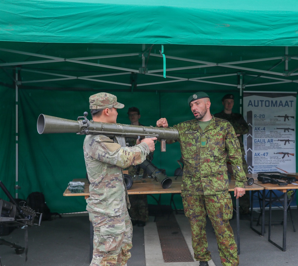 Two military personnel are at an outdoor weapons display booth under a green canopy tent. One soldier in US Army OCP (Operational Camouflage Pattern) uniform is handling what appears to be an RPG-7 or similar rocket-propelled grenade launcher, while a second soldier in a different national camouflage uniform and green beret provides instruction or demonstration. A display board in the background labeled 'AUTOMAAT' lists various firearms including R-20, AK-4, Galil AR, AK-47, and Mosin-Nagant M1891.