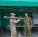 Two military personnel are at an outdoor weapons display booth under a green canopy tent. One soldier in US Army OCP (Operational Camouflage Pattern) uniform is handling what appears to be an RPG-7 or similar rocket-propelled grenade launcher, while a second soldier in a different national camouflage uniform and green beret provides instruction or demonstration. A display board in the background labeled 'AUTOMAAT' lists various firearms including R-20, AK-4, Galil AR, AK-47, and Mosin-Nagant M1891.