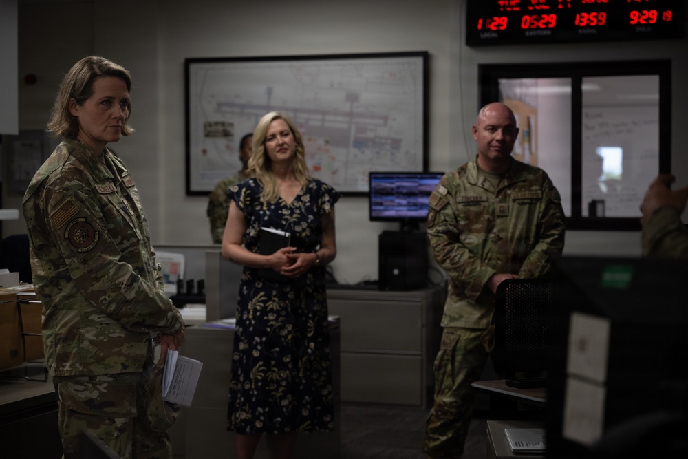 The image shows a military operations or command center environment with two uniformed personnel in camouflage (OCP/MultiCam) pattern and one civilian woman in a floral dress. The group appears to be engaged in a briefing or tour of the facility. Multiple digital clocks displaying different time zones are visible on the wall in the background.