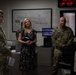 The image shows a military operations or command center environment with two uniformed personnel in camouflage (OCP/MultiCam) pattern and one civilian woman in a floral dress. The group appears to be engaged in a briefing or tour of the facility. Multiple digital clocks displaying different time zones are visible on the wall in the background.