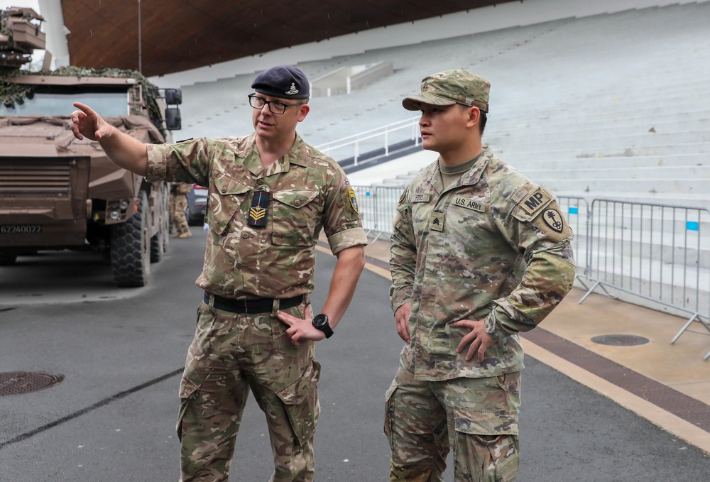 Two military personnel from different nations are engaged in conversation near military vehicles at what appears to be a large venue or stadium. The soldier on the left wears British Army uniform with a dark beret and sergeant chevrons, while the soldier on the right wears U.S. Army Military Police (MP) uniform. The British soldier is pointing in a direction, suggesting he is briefing or orienting the U.S. Army MP sergeant.