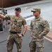 Two military personnel from different nations are engaged in conversation near military vehicles at what appears to be a large venue or stadium. The soldier on the left wears British Army uniform with a dark beret and sergeant chevrons, while the soldier on the right wears U.S. Army Military Police (MP) uniform. The British soldier is pointing in a direction, suggesting he is briefing or orienting the U.S. Army MP sergeant.