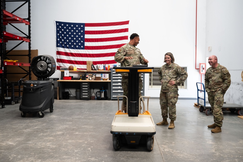 Three military personnel in OCP (Operational Camouflage Pattern) uniforms are gathered in what appears to be a military warehouse or maintenance facility. One service member stands behind a yellow electric utility cart/vehicle while two others stand nearby, all appearing to be engaged in a casual, positive interaction. A large American flag is prominently displayed on the wall behind them.