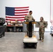 Three military personnel in OCP (Operational Camouflage Pattern) uniforms are gathered in what appears to be a military warehouse or maintenance facility. One service member stands behind a yellow electric utility cart/vehicle while two others stand nearby, all appearing to be engaged in a casual, positive interaction. A large American flag is prominently displayed on the wall behind them.