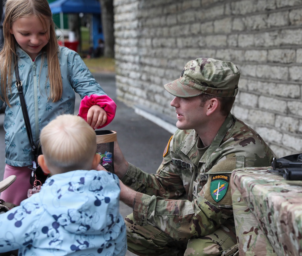 A U.S. Army soldier in OCP (Operational Camouflage Pattern) uniform is crouching down to interact with two young children at what appears to be a community or public outreach event. The soldier is holding a container, possibly offering items to the children. The setting appears to be outdoors near a stone or brick wall.