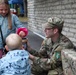A U.S. Army soldier in OCP (Operational Camouflage Pattern) uniform is crouching down to interact with two young children at what appears to be a community or public outreach event. The soldier is holding a container, possibly offering items to the children. The setting appears to be outdoors near a stone or brick wall.
