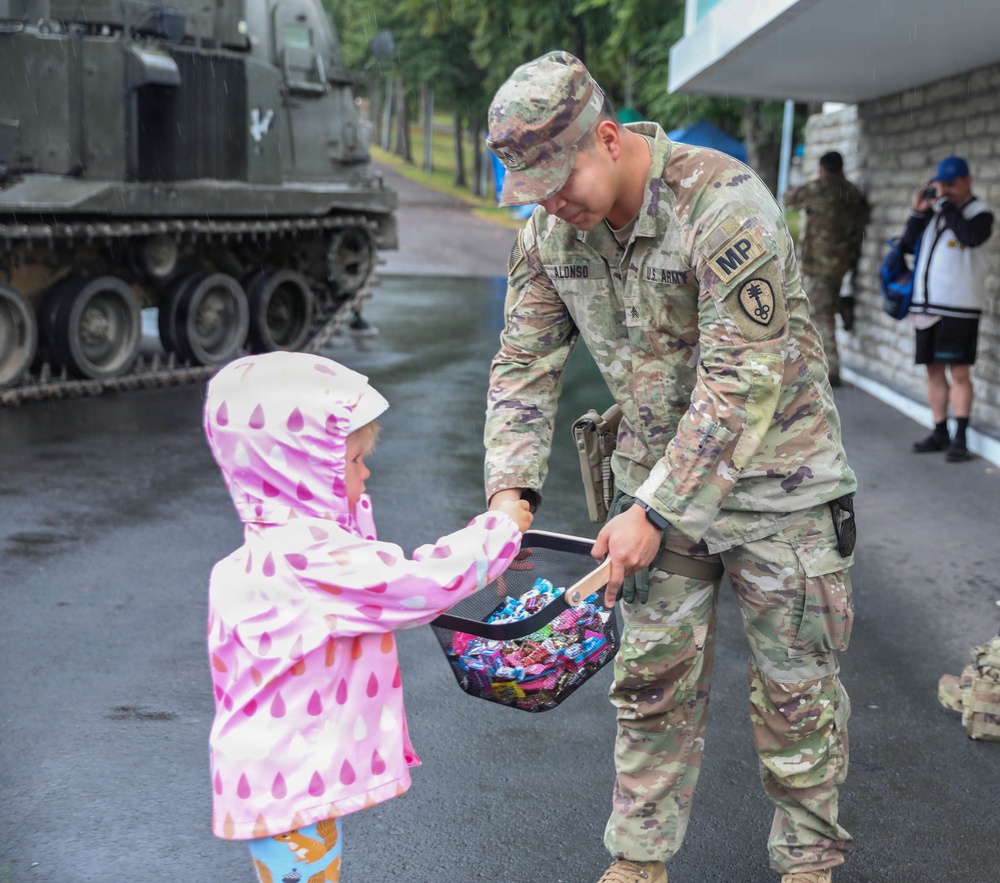 A U.S. Army Military Police soldier named Alonso bends down to offer candy from a basket to a young child wearing a pink polka-dot raincoat. The interaction takes place outdoors on a wet surface during rain, suggesting a public military event or community engagement activity. A military armored vehicle is visible in the background along with a civilian photographer.