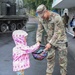 A U.S. Army Military Police soldier named Alonso bends down to offer candy from a basket to a young child wearing a pink polka-dot raincoat. The interaction takes place outdoors on a wet surface during rain, suggesting a public military event or community engagement activity. A military armored vehicle is visible in the background along with a civilian photographer.